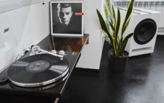 A close-up picture of a vinyl player. Beside the player, there is a Kraftwerk album, a handcrafted stereo from Italy, with a beautiful plant in the background.