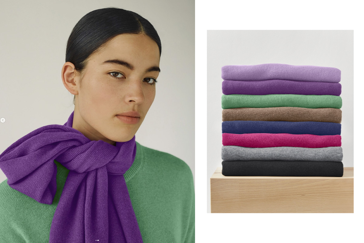 A female model wearing a green hessnatur cashmere sweater and a purple scarf stands beside a stack of neatly folded cashmere sweaters in a variety of colors.