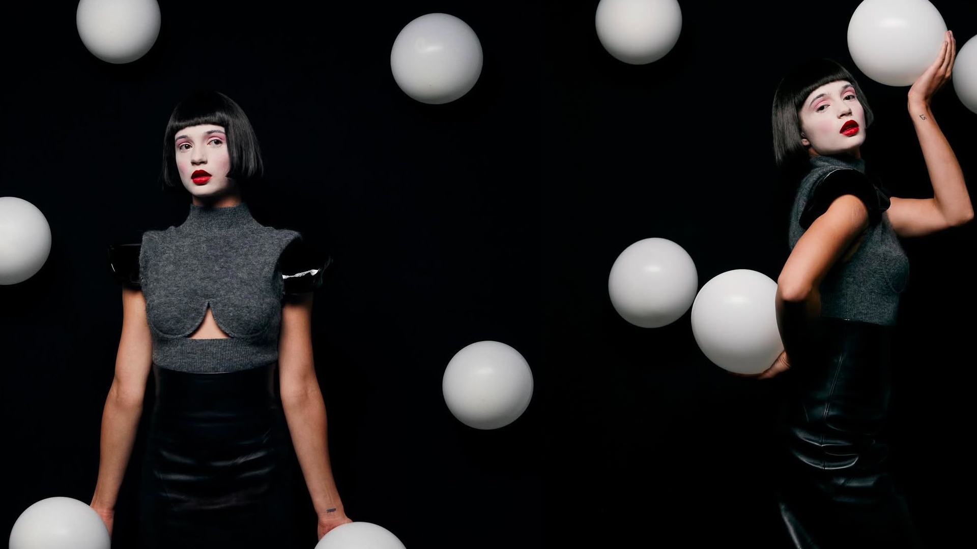 Lisa-Marie Koroll in a dramatic studio setting, wearing a futuristic black and grey ensemble. White spherical props float around her against a dark background, adding a surreal, artistic effect.