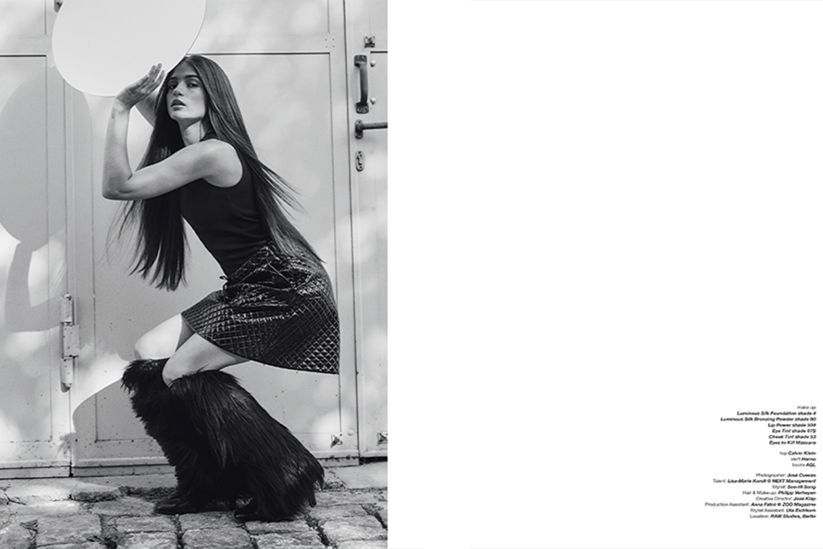 A black-and-white photograph of Lisa-Marie Koroll posing outdoors. She kneels gracefully on cobblestone, wearing a textured skirt, fur boots, and a sleeveless top, with her long hair cascading down her back.