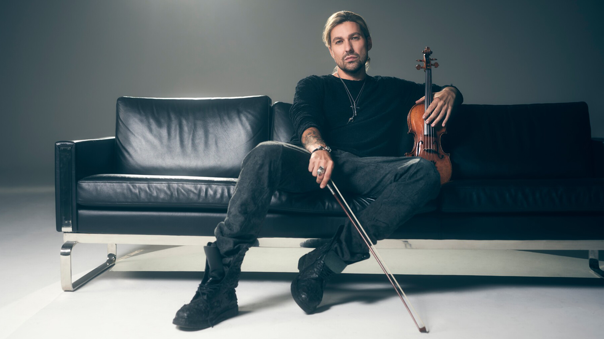 David Garrett, dressed in black, sits on a sleek black leather couch holding his violin. His relaxed pose, intense gaze, and dimly lit studio setting create a moody, sophisticated atmosphere.