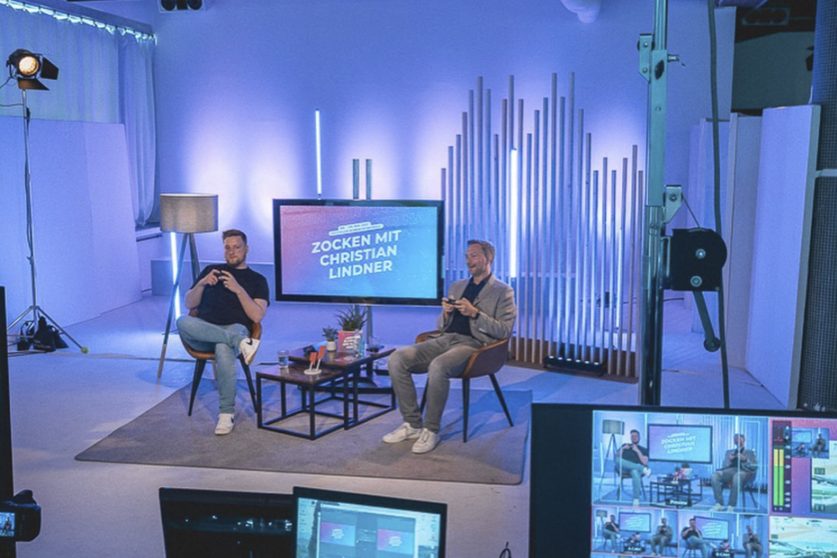 A modern talk show-style setup at raw studios., featuring two speakers engaged in a gaming session. A large screen behind them displays “Zocken mit Christian Lindner,” with cool-toned lighting and a contemporary backdrop enhancing the setting.