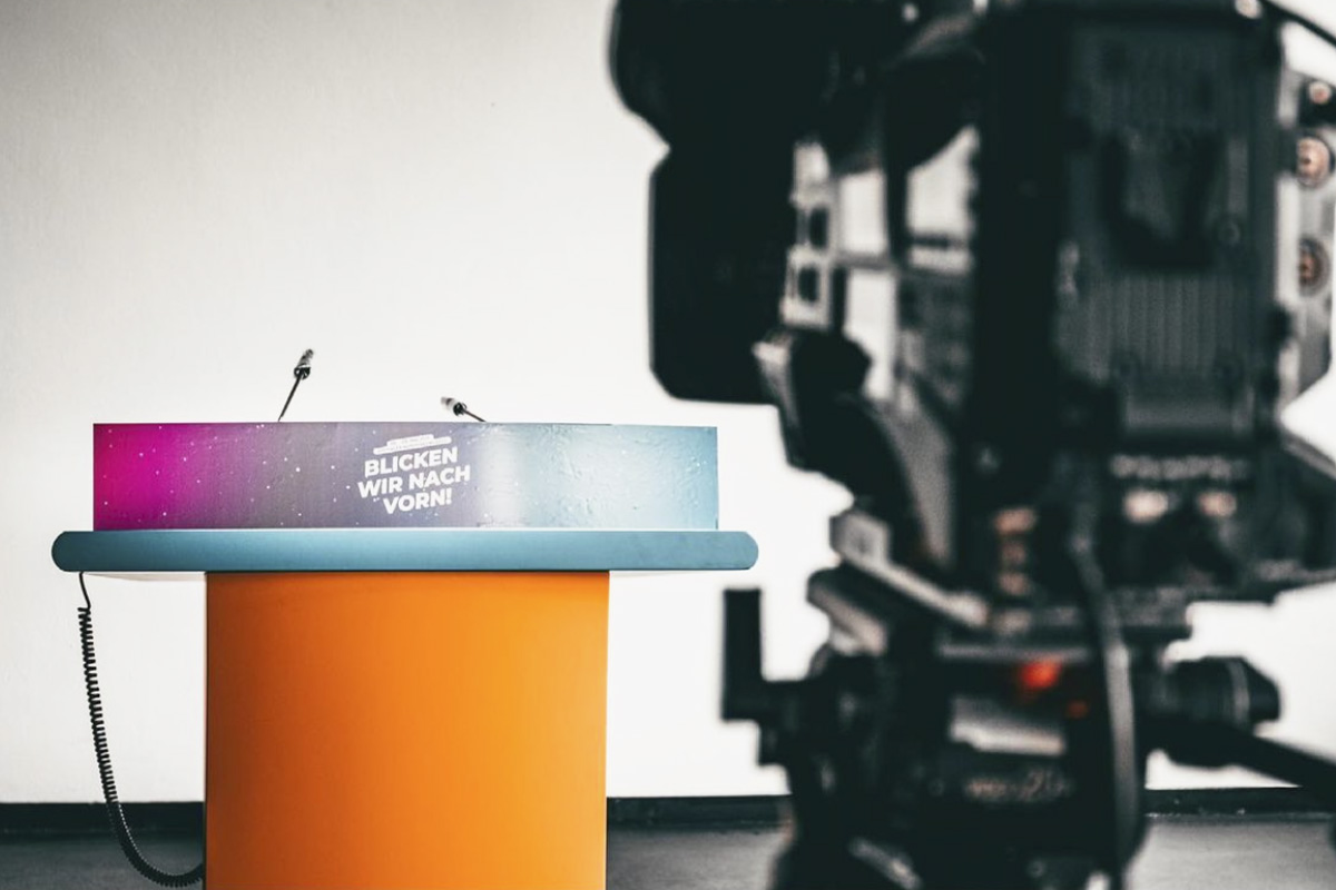 A close-up of a stage podium at raw studios. with the phrase “Blicken wir nach vorn!” printed on the front. A professional camera in the foreground is focused on the stage setup, ready for the event broadcast.