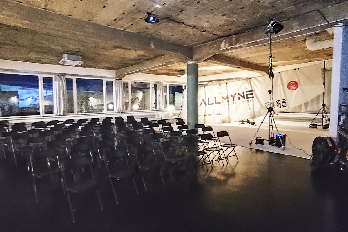 An intimate event setup at raw studios. with rows of black chairs facing a stage area. A large sail with the "ALLMYNE" logo serves as a backdrop, while a projector screen displays visuals related to the Ocean Race.