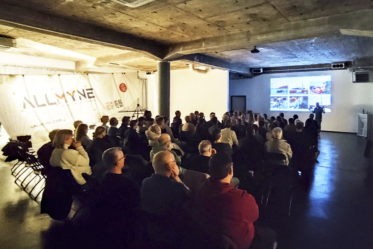 A captivated audience at raw studios. attentively listening to Robert Stanjek as he presents insights from his Ocean Race experience. The projection screen displays dynamic images related to the race