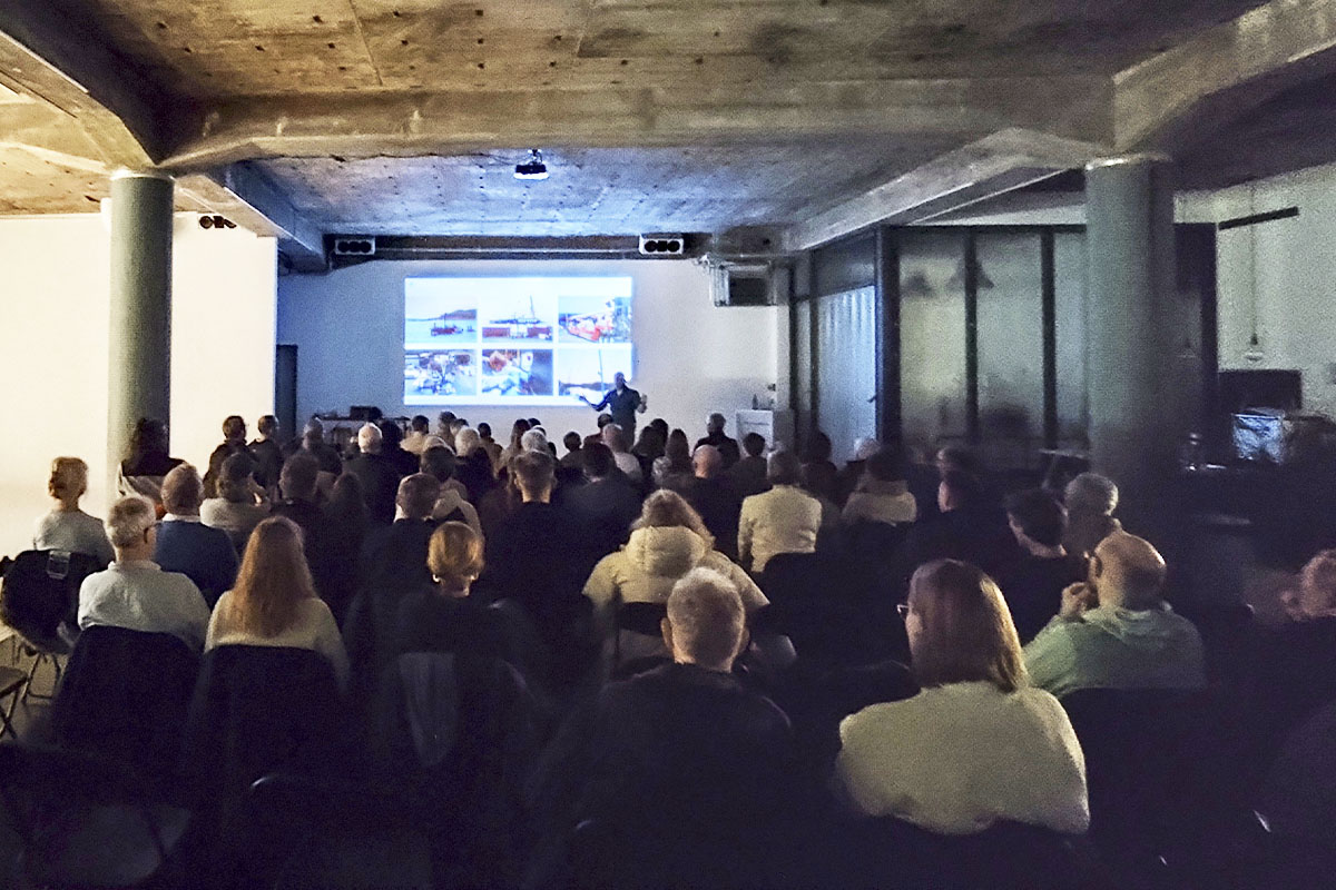 A speaker on stage at raw studios. engaging a full audience, with visuals from the Ocean Race projected onto the wall. The minimalist, industrial-style venue creates a focused and professional atmosphere.