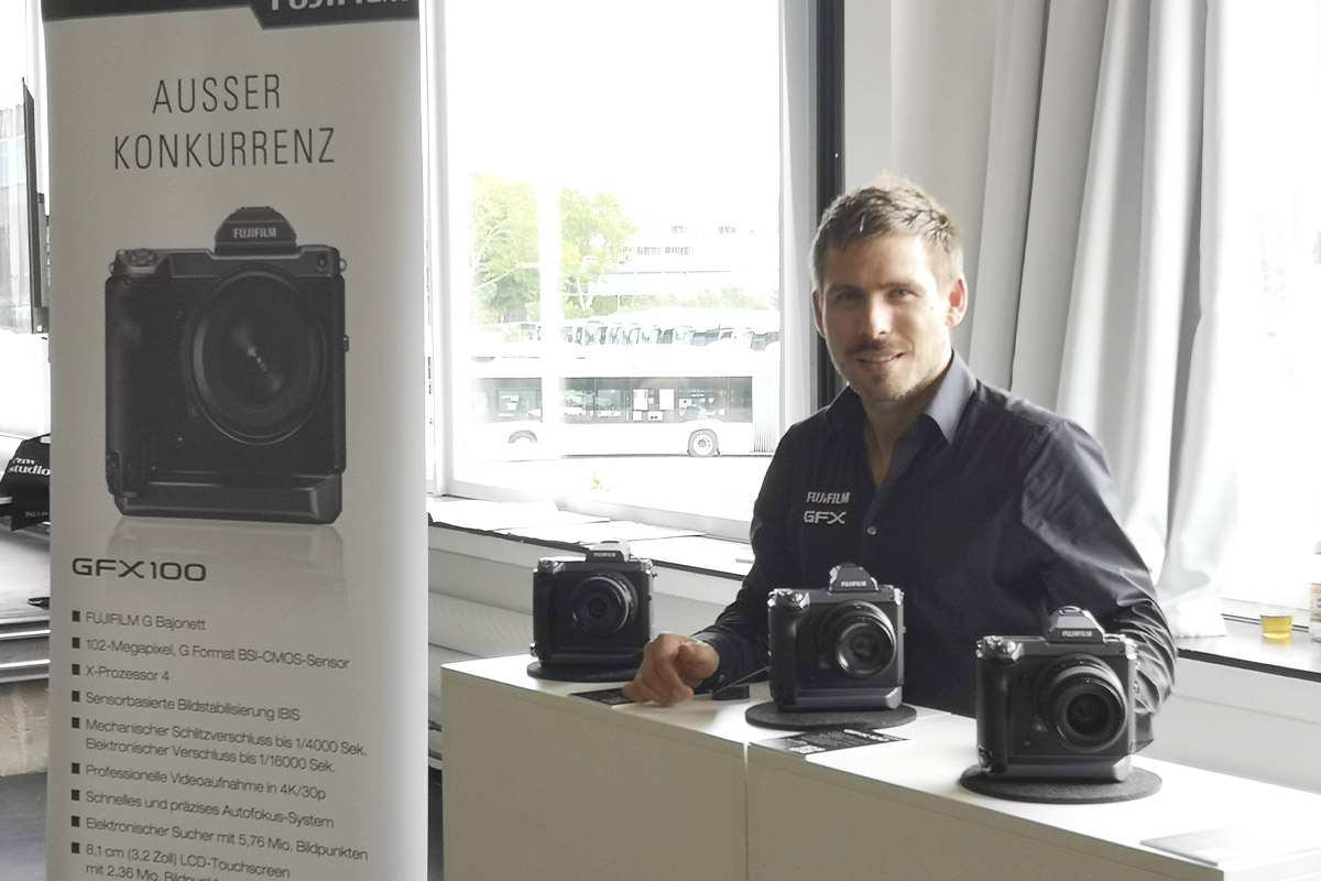 A Fujifilm representative standing behind a display table with multiple Fujifilm GFX100 cameras, engaging with attendees at the Fuji Road Show at raw studios.