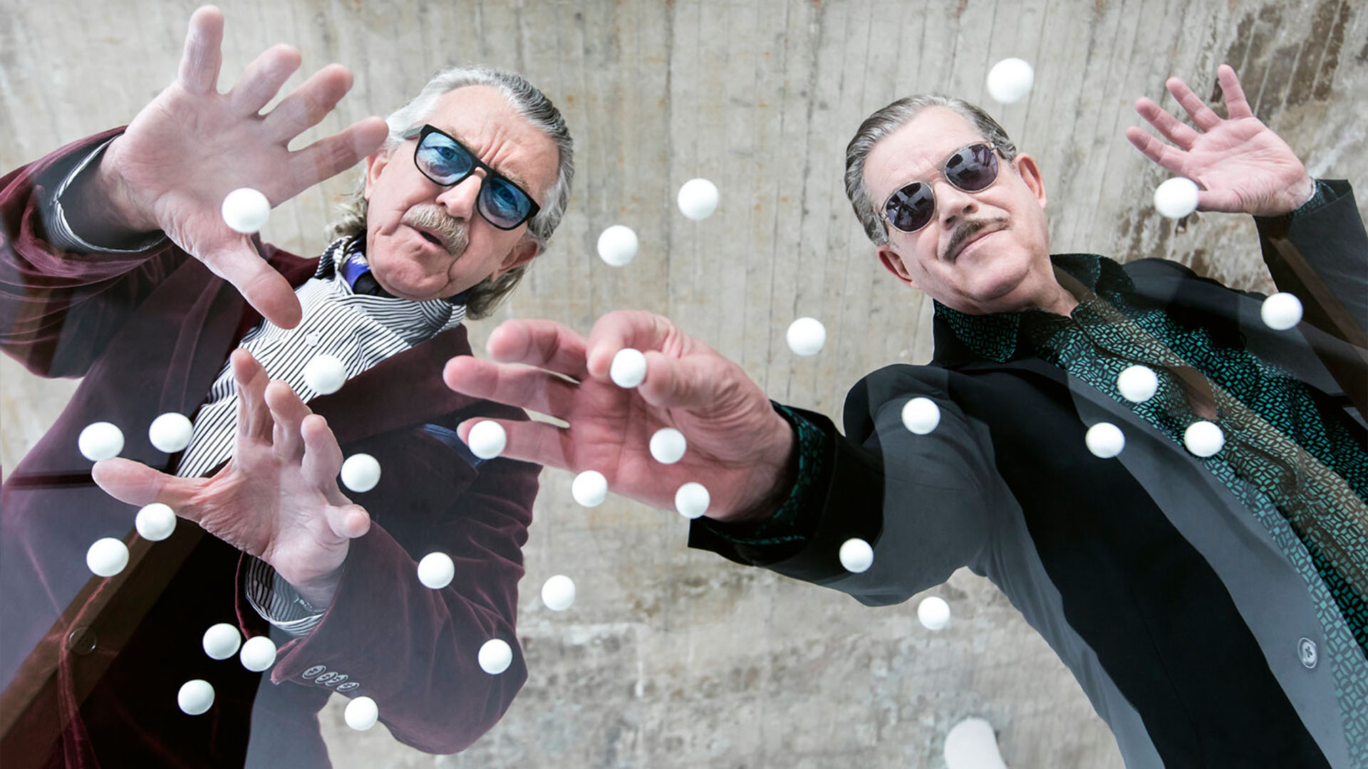 The legendary electronic duo YELLO, dressed in stylish suits and sunglasses, are seen from a low-angle perspective, reaching out towards floating white spheres. Their playful expressions and the creative composition add a surreal, futuristic touch.