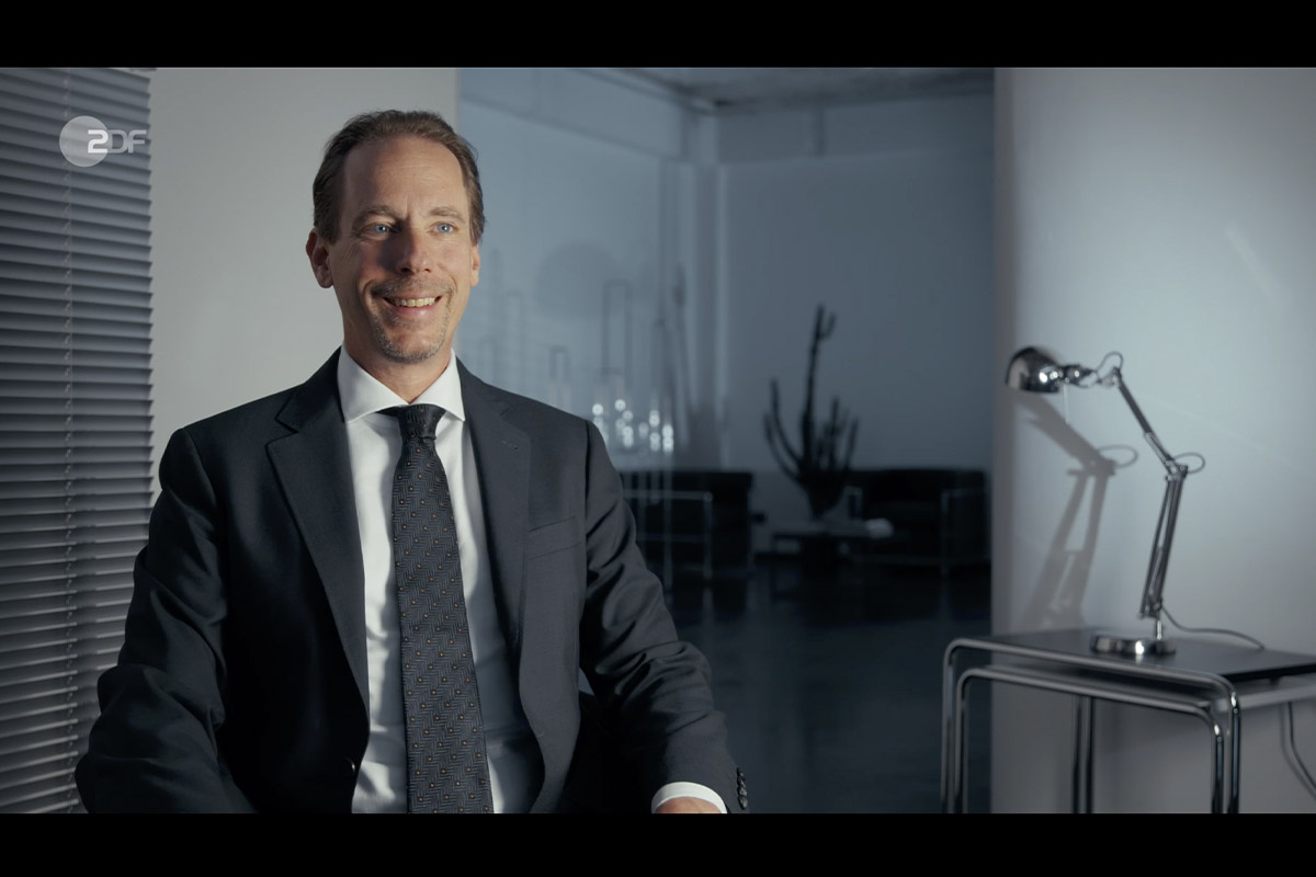 Interview shot of Alexander Heist in a suit, seated in a modern studio setting with minimalist decor and soft daylight, filmed for the Cum-Ex scandal documentary.
